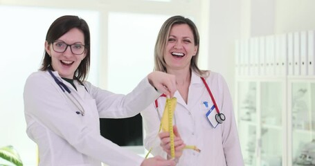 Happy blonde and brunette doctors playfully measure banana with measuring tape in clinic. Cheerful women medics in uniform enjoy break laughing together - Powered by Adobe