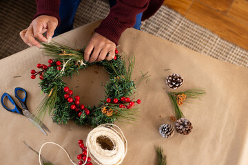 Asian senior woman rafting festive wreath with pine cones and berries at home for christmas