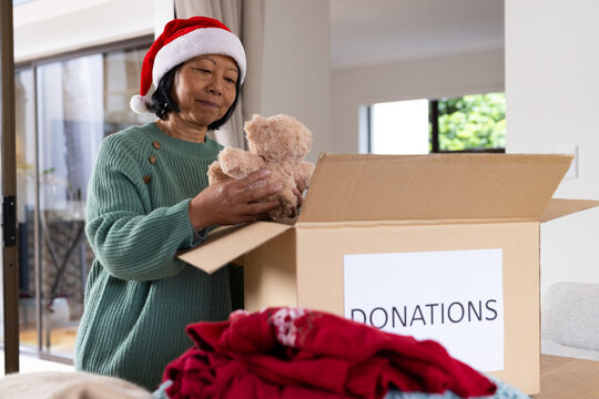 Senior asian senior woman in Santa hat packing teddy bear into donation box at home, Christmas time - Powered by Adobe