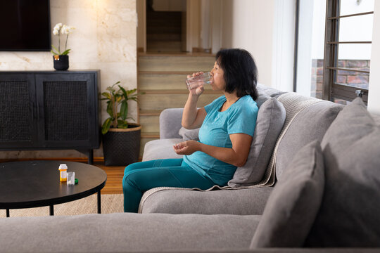 Asian senior woman sitting on sofa drinking water, taking medication at home