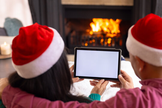 Chrsitmas time, multiracial senior couple in santa hats using tablet at home, copy space