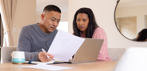 Multiracial senior couple reviewing documents on laptop together at home, focused and engaged