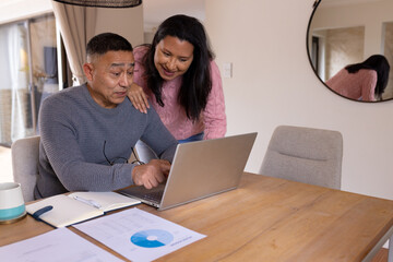 Multiracial senior couple using laptop at home, discussing finances with documents on table