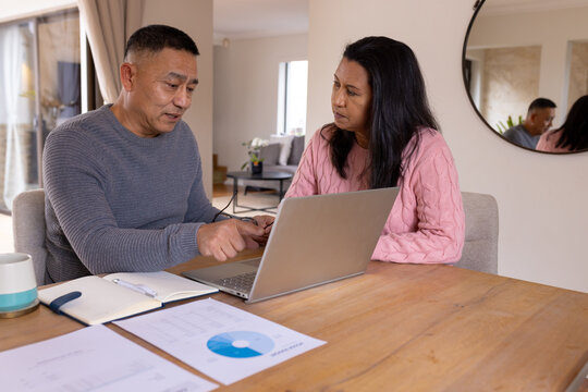 Multiracial senior couple discussing finances at home using laptop and documents on table