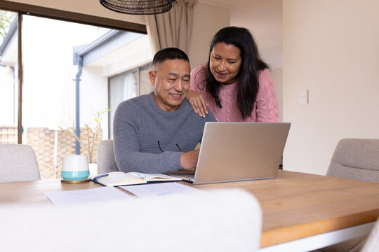 Multiracial senior couple smiling while using laptop at home, planning holiday together