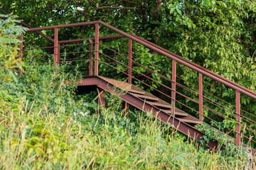 Fototapeta premium An old red metal staircase with railings surrounded by lush green foliage, viewed from below under a hill. The weathered structure blends into nature under soft daylight during a summer day outdoors. 
