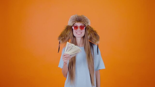 A girl in funny pink glasses and a ushanka hat playfully holds a fan of American dollar bills, showcasing her fun personality. The vibrant orange background enhances the playful atmosphere.