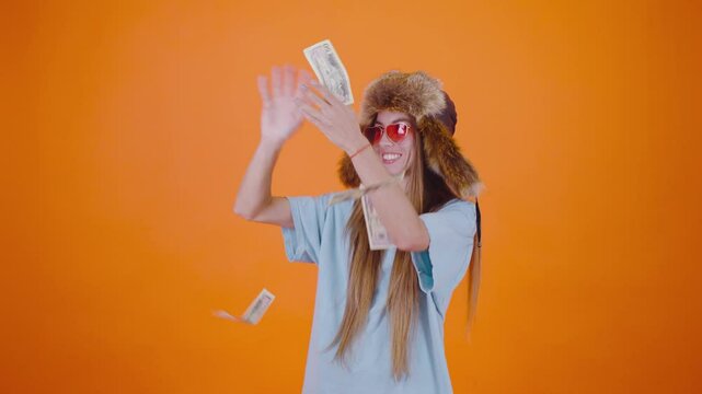 A girl in funny pink glasses and a ushanka hat joyfully throws a large amount of money into the air, showcasing her playful expression of wealth and abundance. The vibrant orange background adds to