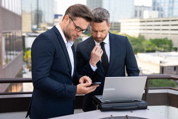 Two businessman on the street, deeply engrossed, eyes fixed on their laptop screen. Two handsome...