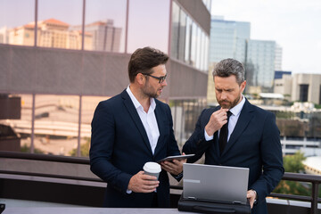 Two businessman on the street, deeply engrossed, eyes fixed on their laptop screen. Two handsome...