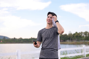 Smiling young sport man wiping sweat with towel while resting after a running by the lake