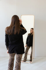 Back of young woman standing in front of mirror in black blazer and leopard print pants poses confidently in minimalist modern interior studio. Female model in fashion-forward outfit, elegant style
