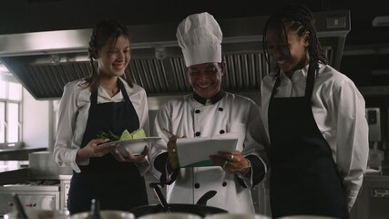 Professional Asian senior male chef in uniform with chef hat standing teaching cooking methods to his female multiracial students listening intently and taking notes in the kitchen at the restaurant.