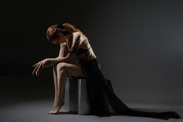 A contemplative dancer in a flowing dress poses on a stool in dim lighting © Wisky