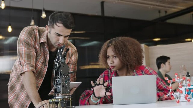 Cute African American female student with an afro hairstyle and her Caucasian male teacher use laptop and tablet with a program while learning future technology robotic hand and arm in the classroom. 