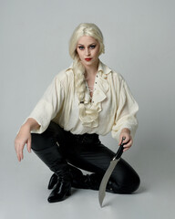 full length portrait of beautiful female model, wearing vintage gothic fantasy ruffle blouse white shirt, leather pants, blonde hair. holding knife weapon. sitting pose, isolated on studio background.