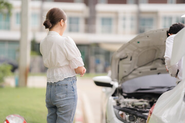 Car Trouble: A woman anxiously looks on as a man inspects the engine of a broken down car. The...
