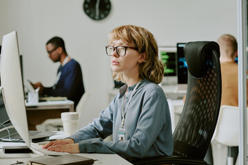 Medium shot of young Caucasian female specialist sitting at workplace in modern office looking...