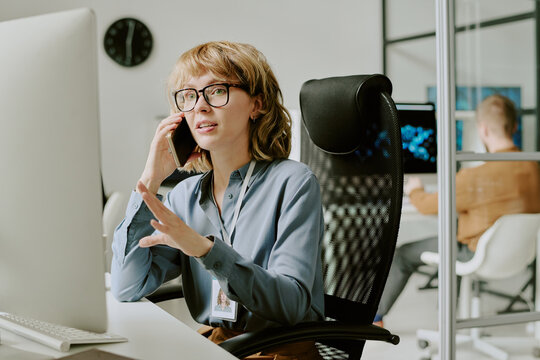 Medium shot of young woman wearing eyeglasses sitting at workplace in modern office looking at computer screen and talking on phone