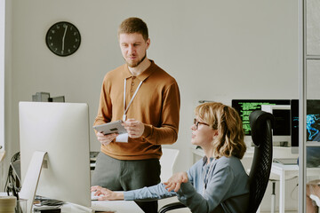 Young Caucasian man with digital tablet in hands and woman sitting at office desk talking about new...