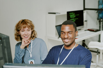 High angle view of two cheerful young Black and Caucasian specialists smiling while working together on computer in IT company office