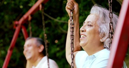 Indian Asian mid age couple in their fifties with gray hair sitting on swings in the park, smiling at the camera, enjoying a peaceful moment outdoors in daylight
