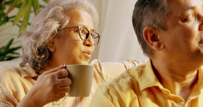 Indian Asian couple in their fifties with gray hair enjoying tea or coffee by a sunny window, relaxing on the sofa and spending quality time together in the morning