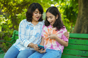 Young Indian mother and daughter using smart phone at summer public park having fun together. Wireless Technology, 5G Network, Video Entertainment. Social media.