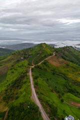 Aerial shot of a winding road through lush green hills with a cloudy sky, capturing the essence of a tranquil mountain landscape.