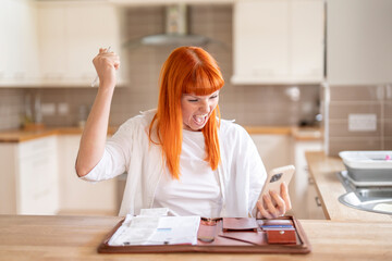 Excited woman celebrating financial victory while managing bills in a bright kitchen on a sunny day