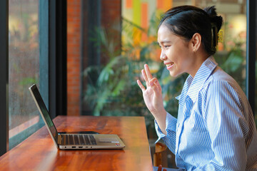 excited asian woman sitting in cafe looking at laptop on wooden table have online meeting, video chat, showing ok signs wearing striped blue shirt