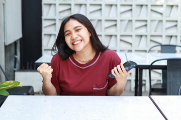 pretty asian woman celebrate winning playing online game tournament using smartphone with raising fist say yes gesture sitting in outdoor cafe