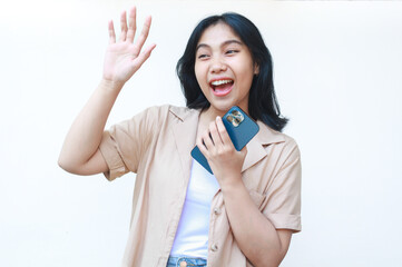 happy asian woman holding smartphone looking away with wave hand wearing casual brown clothes isolated on white background