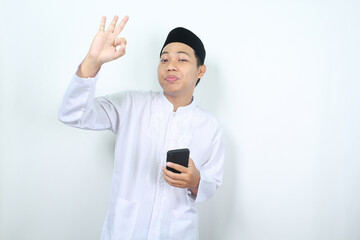 smiling asian muslim man showing okay signs with holding mobile phone isolated