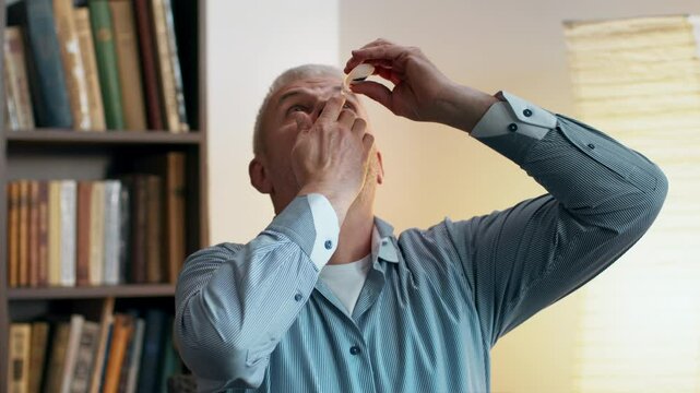 A man uses eye drops while seated at a desk, surrounded by shelves filled with books, ready for online class