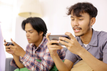 Two Young Asian Men Wearing Casual Clothes Together Playing Video Games Online On Mobile Phone At Home