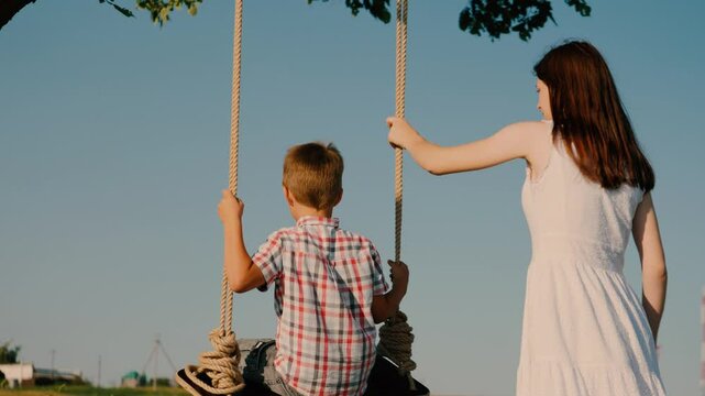 Attractive mother gently pushes junior son on swing spending time in nature. Mom and son spend quality time together in backyard rocking on swing. Child enjoys wonderful time with parent on swing