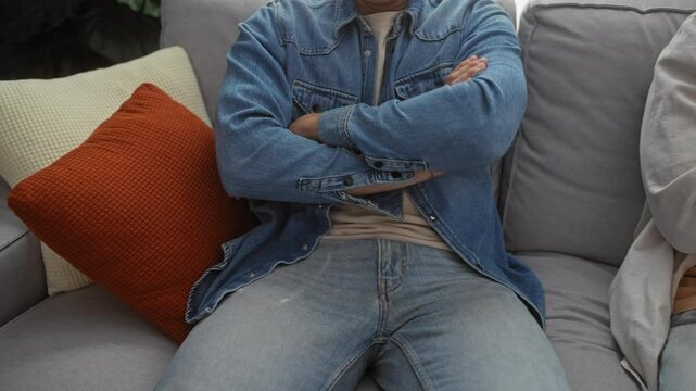 Men sitting on a sofa in a living room wearing casual clothes with crossed arms and jeans in an indoor home setting