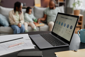 Family sitting on couch engaging in financial planning activity visible on laptop screen on table foreground. Parents and kids interacting, discussing important financial information