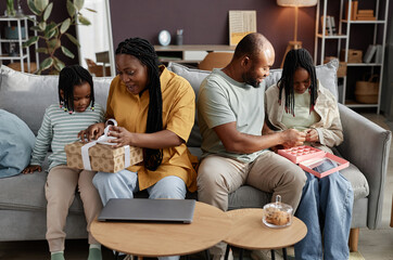 Family enjoying time together on couch while unwrapping gifts and interacting with tablet in cozy living room, displaying connection and happiness