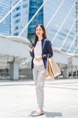 Fototapeta premium Vertical portrait asian business woman standing at outdoor with shopping bags city building background