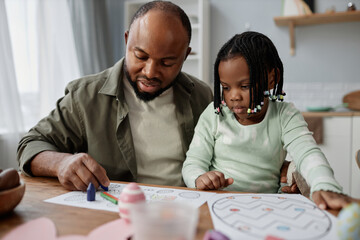 African American father sitting with daughter, both focusing intently on homework spread out on table in cozy kitchen setting. Daughter holding crayon, father guiding her learning activity