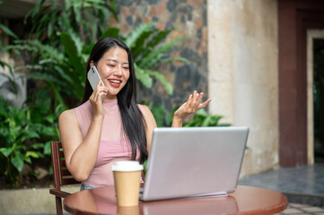 An attractive Asian businesswoman is talking on the phone while looking at her laptop screen.