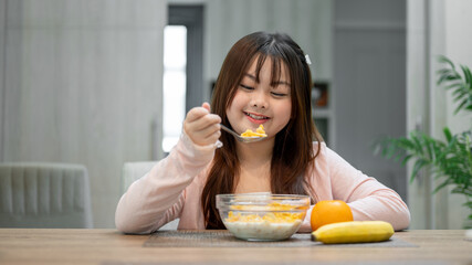 A young, cute Asian girl is having breakfast, enjoying eating a bowl of cereal at a dining table.