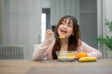 A young, cute Asian girl is having breakfast, enjoying eating a bowl of cereal at a dining table.