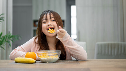 A young, cute Asian girl is having breakfast, enjoying eating a bowl of cereal at a dining table.