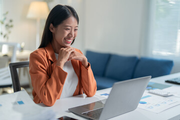 Asian businesswoman happily works from her home office, smiling at her laptop screen while tackling tasks related to business, finance, and sales