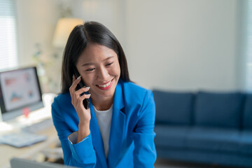 Young asian businesswoman happily works in her home office, smiling as she chats on her cell phone, surrounded by technology and a laptop on her desk