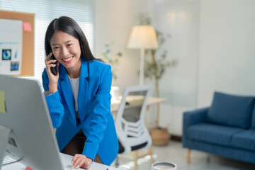 Obraz premium Young asian businesswoman multitasks happily in a modern office, talking on her phone and working on her computer, exuding confidence and professionalism
