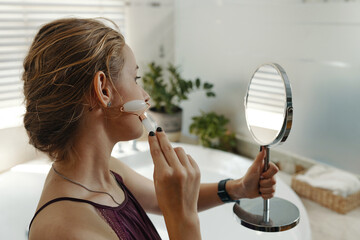 A woman in casual attire applying face skincare with a brush, holding a handheld mirror. Indoor background features natural light and green plants © DragonImages